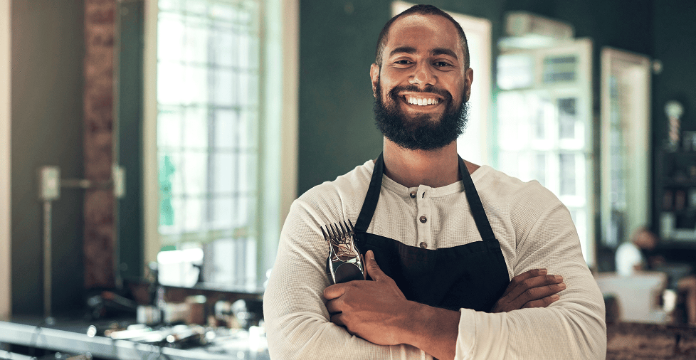 A smiling person in an apron holds a knife, ready to prepare food in a kitchen setting